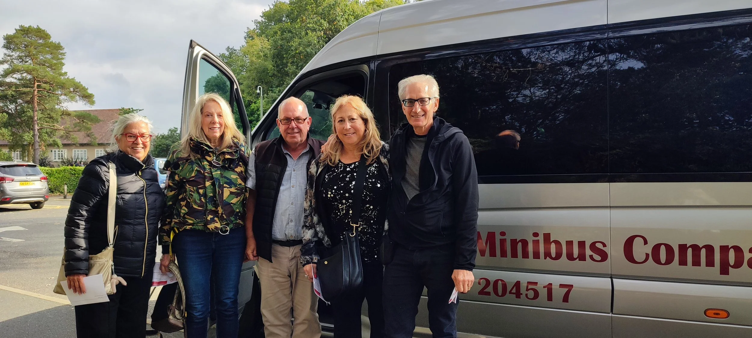 Group of five smiling people standing next to a minibus in a parking lot, with green trees and a building in the background.