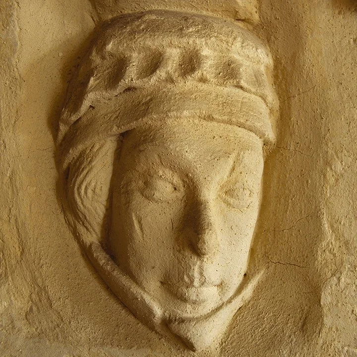 Stone relief sculpture of a woman's face wearing a headscarf or turban, mounted on a textured wall.