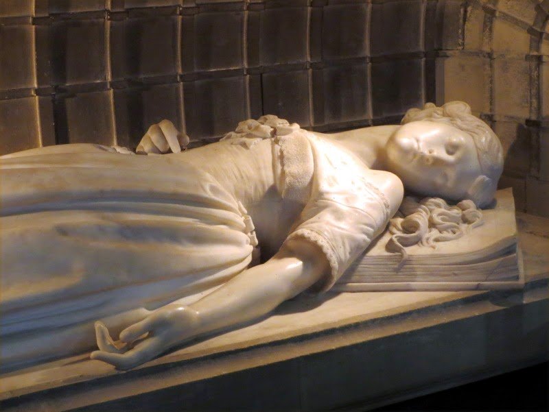 Marble sculpture of a woman lying on a bed with her eyes closed and hair spread out, in a relaxed pose.