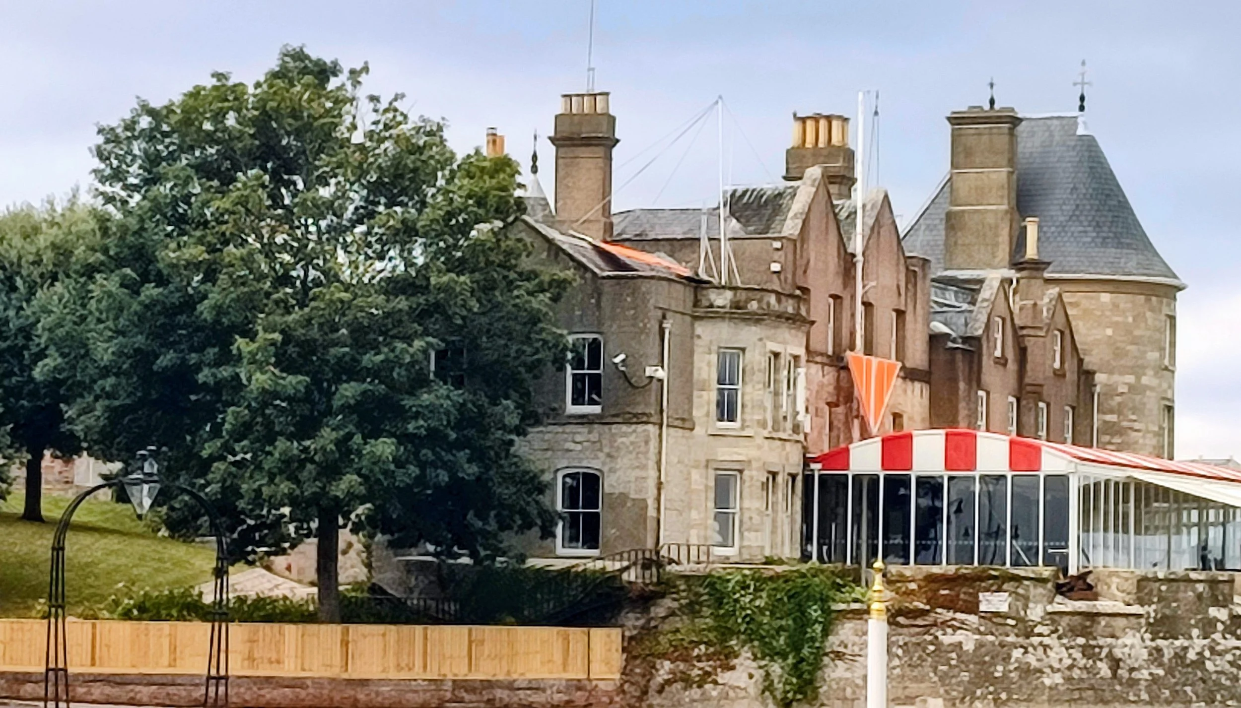 A historic stone castle with multiple towers and chimneys, partially obscured by a large green tree, with an outdoor seating area in the foreground and a cloudy sky in the background.