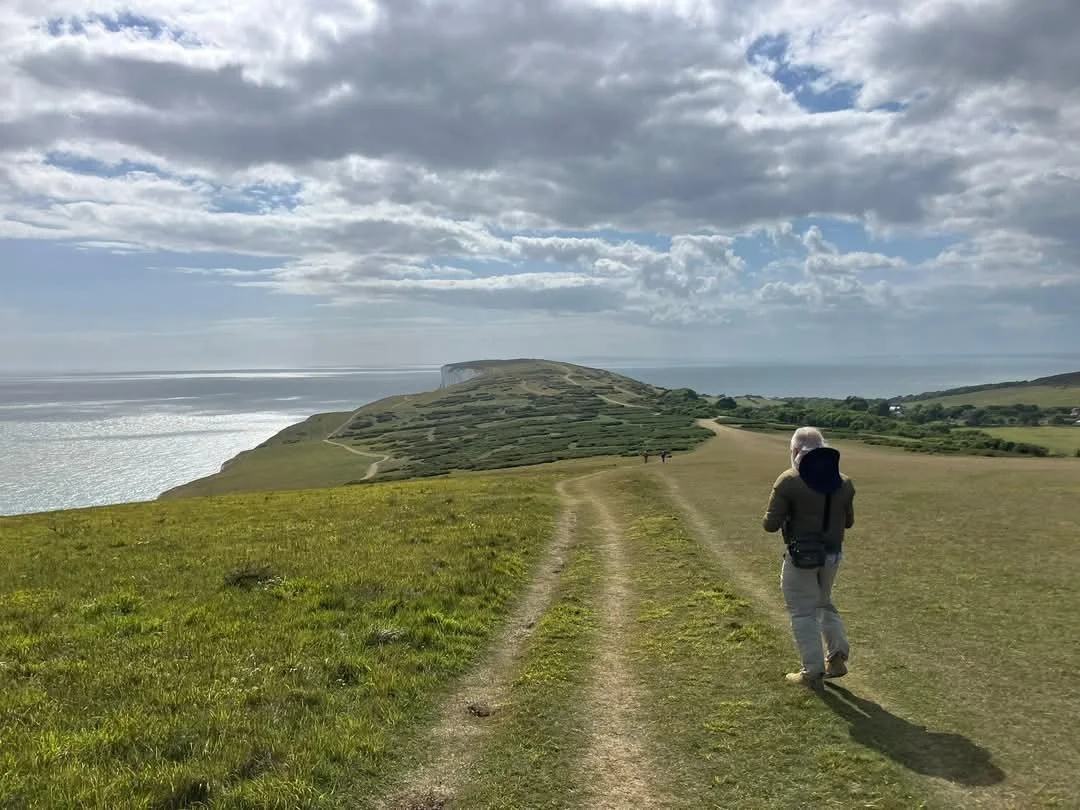 A person walking along a dirt path on a grassy hillside with the ocean in the background, under a partly cloudy sky.