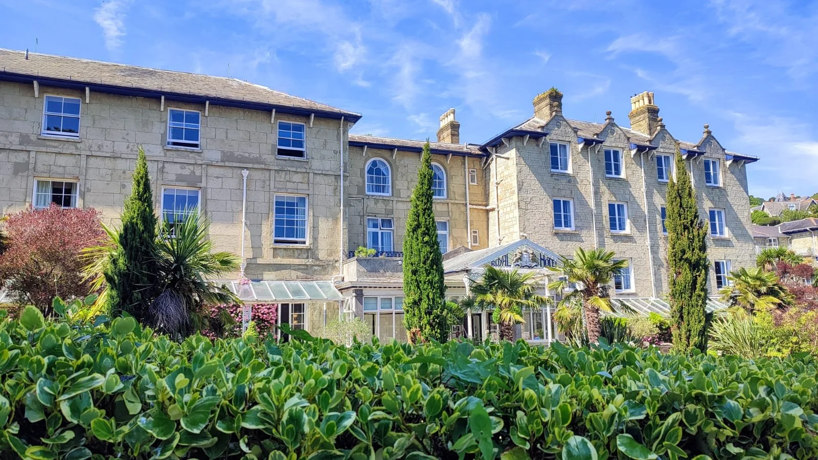 A large, historic stone hotel with a sign reading 'Royal Hotel', surrounded by well-maintained greenery including bushes, palms, and tall cypress trees under a clear blue sky.