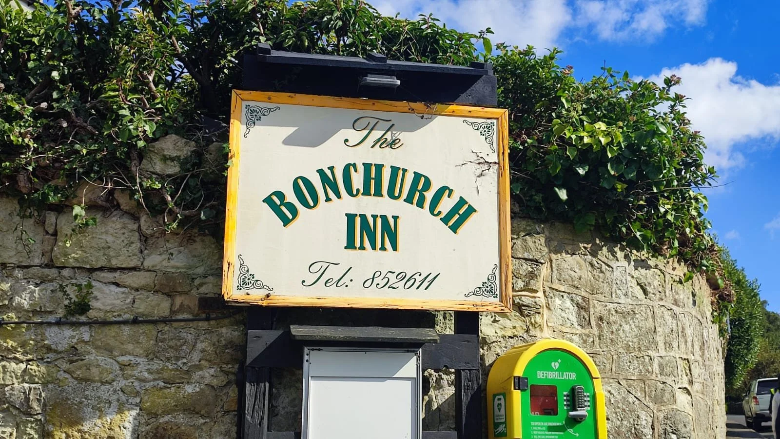 Sign for The Bonchurch Inn with a phone number below, against a stone wall with greenery and a bright blue sky in the background. There is a yellow defibrillator box next to the sign.