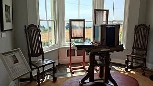 A room with two wooden chairs, a window with natural light, and an artist's easel with canvases, along with a small table and framed pictures.