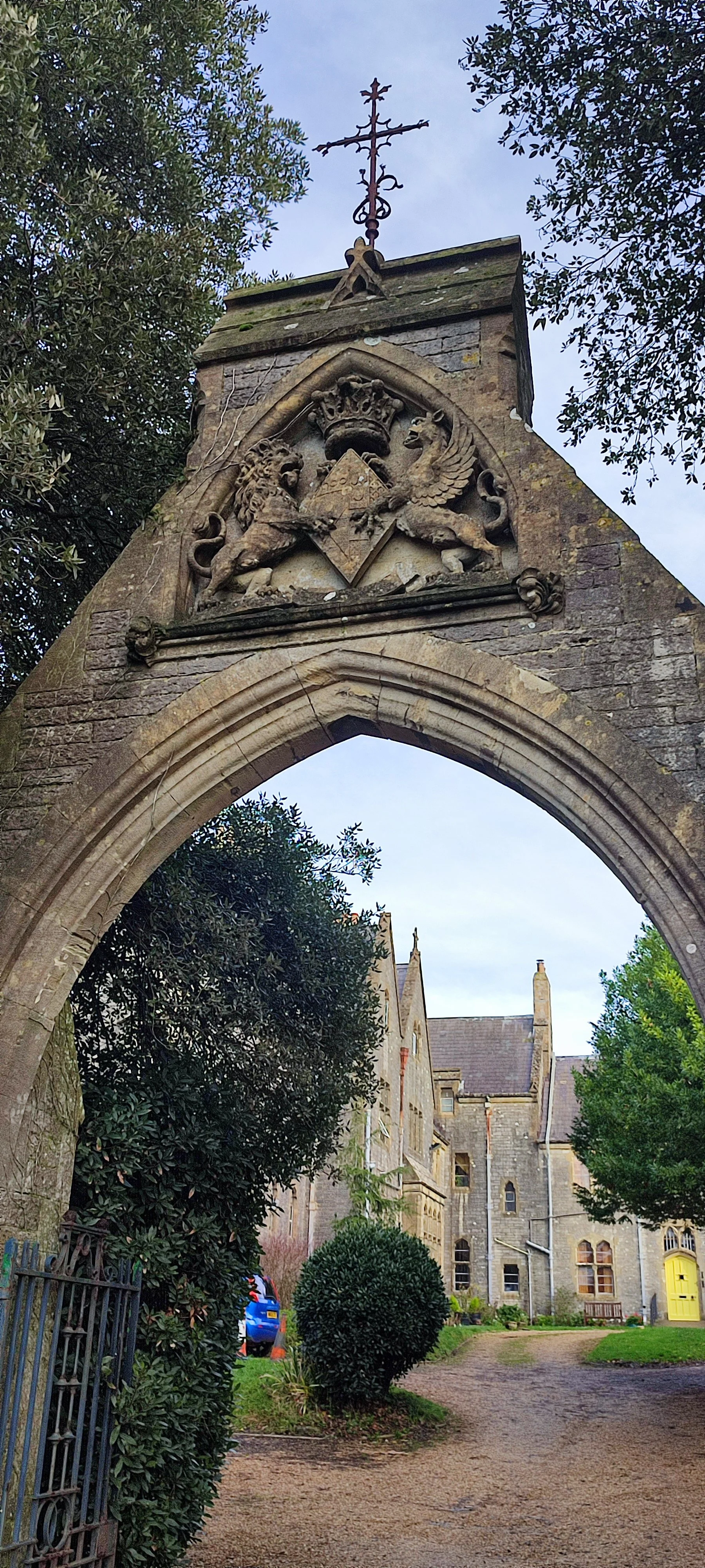 An old stone archway with coat of arms featuring a lion and a griffin, topped with a cross, in front of historic stone buildings and trees.