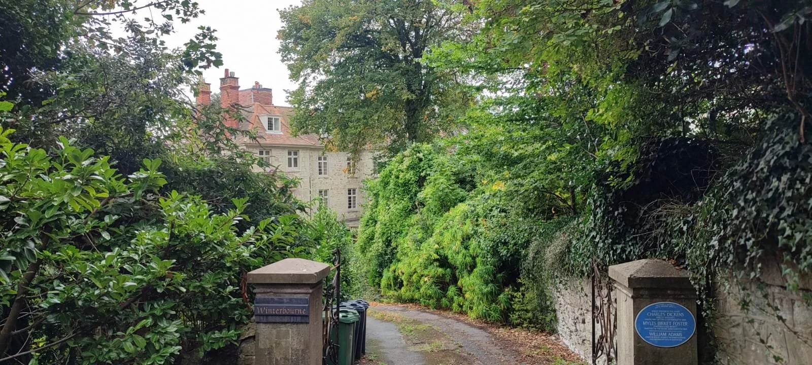 A stone wall and gate at the entrance to a lush green alleyway with a house visible in the background.