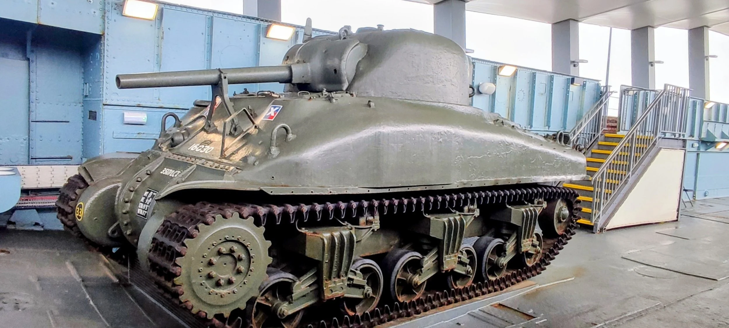 Vintage military tank on display indoors with stairs nearby for viewing, painted in green with markings.