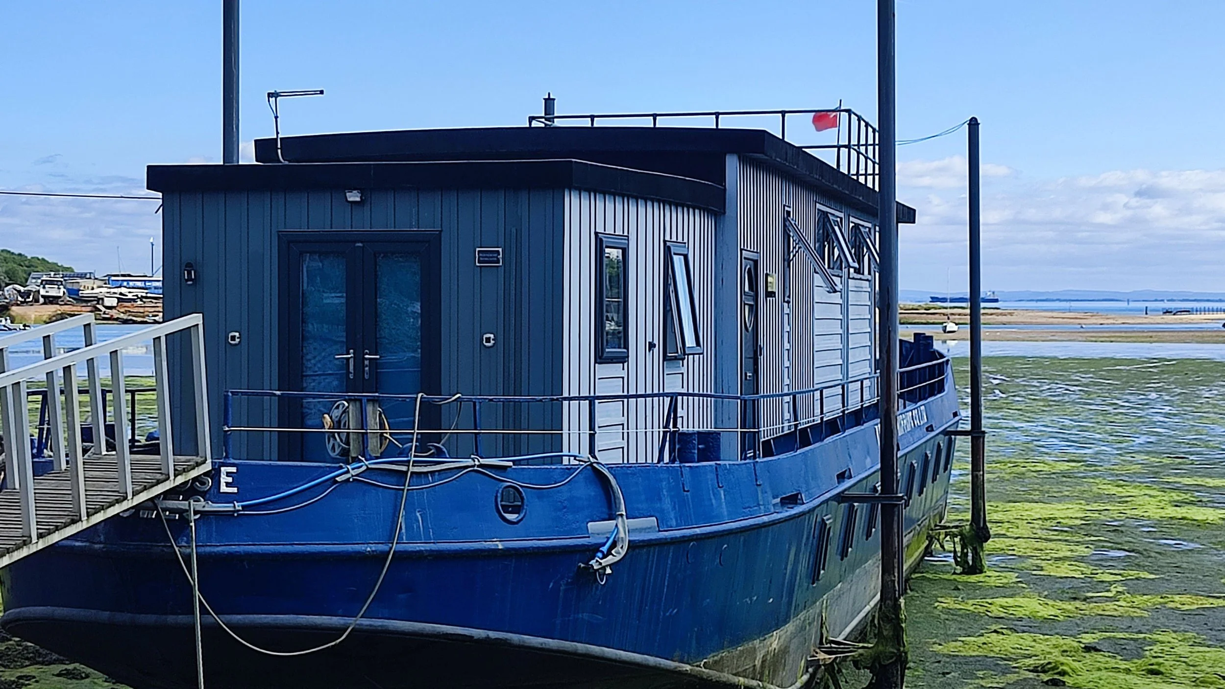 A houseboat with a dark blue exterior is docked at a pier near a body of water, with green algae visible on the shore.