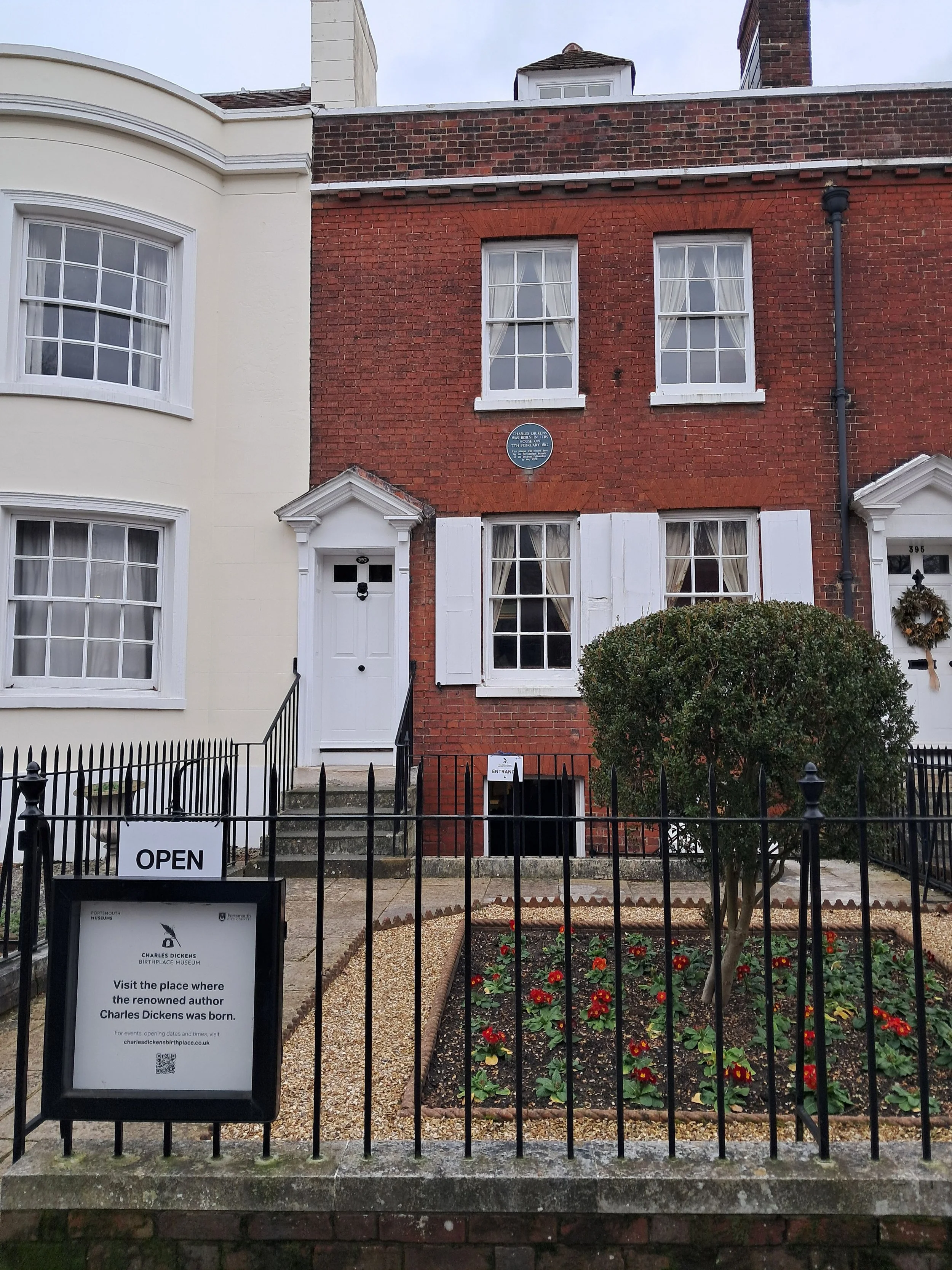 Front view of a house with a small garden in front, black metal fence, sign indicating the Charles Dickens Birthplace Museum, and an 'Open' sign. The house is brick with white window shutters and a white door.