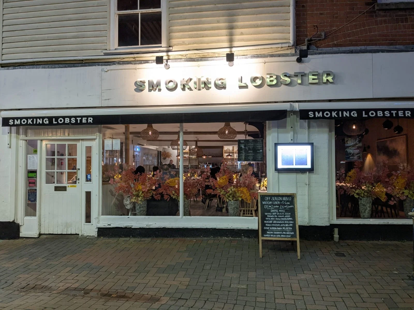 Exterior view of a restaurant named 'Smoking Lobster' with large windows and floral arrangements inside, and a chalkboard menu outside.