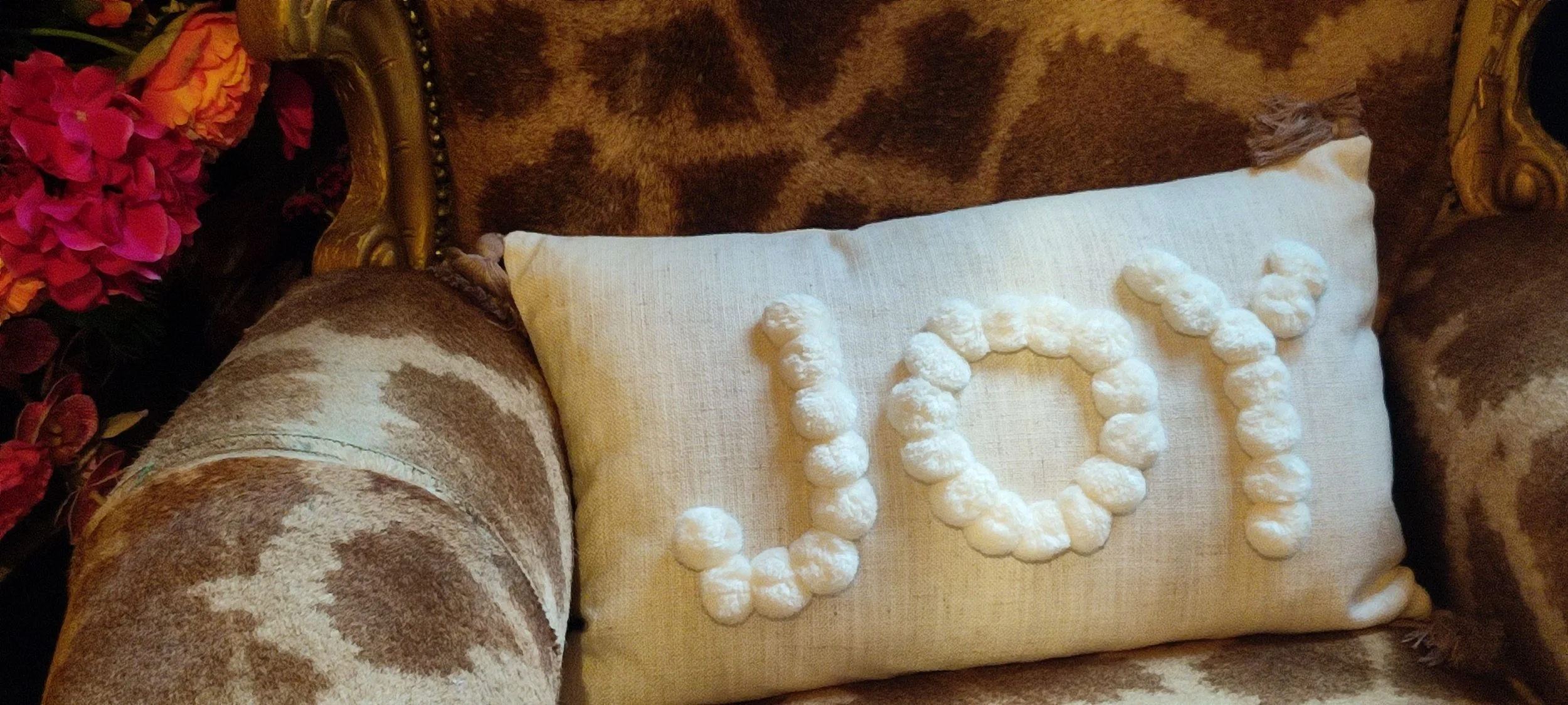 Decorative pillow with white puff pom-poms spelling out "COZY" on a vintage-style sofa with a brown patterned fabric. A floral arrangement with pink and orange flowers is partially visible on the left side.