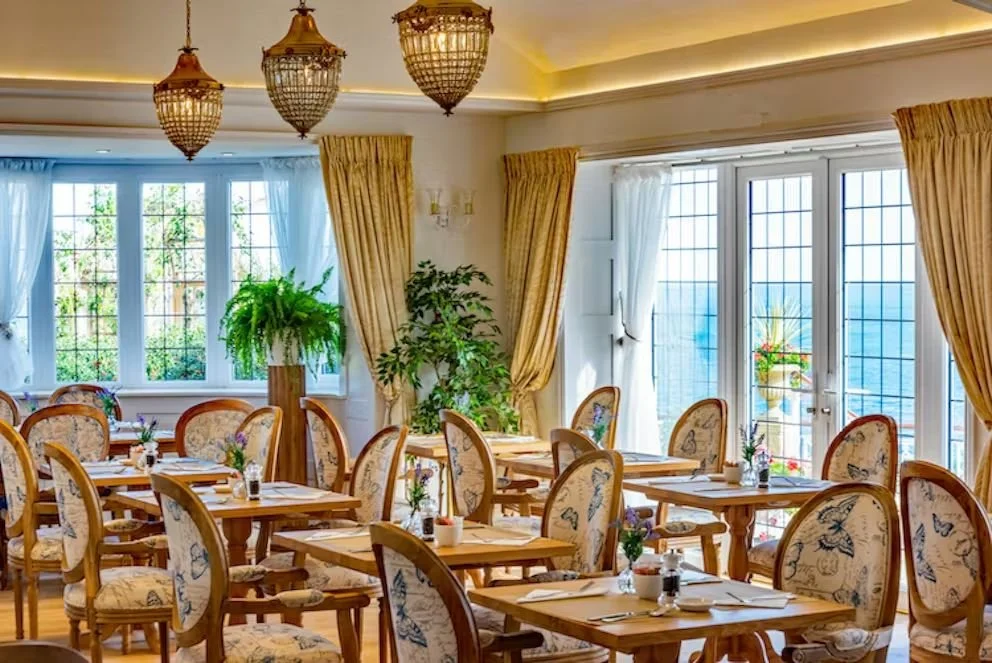 A bright, elegant dining room with large windows, gold curtains, and chandelier lighting. Multiple tables are set with napkins, cutlery, and small flower arrangements. Potted plants add greenery to the space.