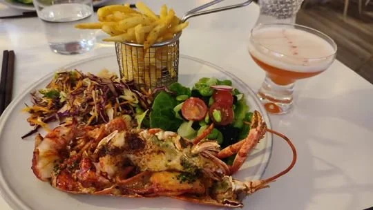 Plate with lobster tail, mixed green salad with cherry tomatoes, and French fries in a small basket, with a glass of beer nearby.