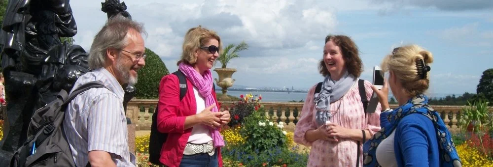 Four women and one man standing outdoors, smiling, with a scenic view in the background, some holding devices, in a garden setting.