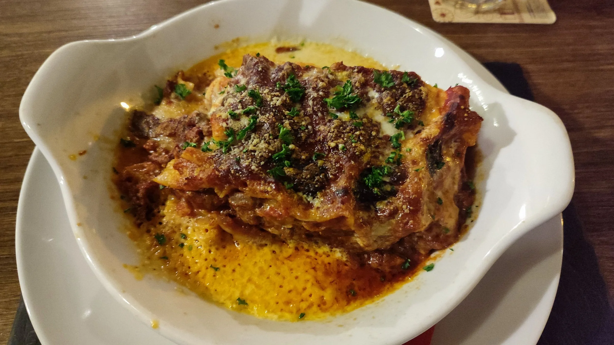 A baked dish with a crispy topping, garnished with chopped parsley, served in a white oval dish on a wooden table.