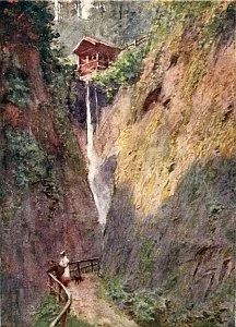 A waterfall flowing down a rocky cliffside with a house at the top, greenery around, and a person walking on a path in the foreground.