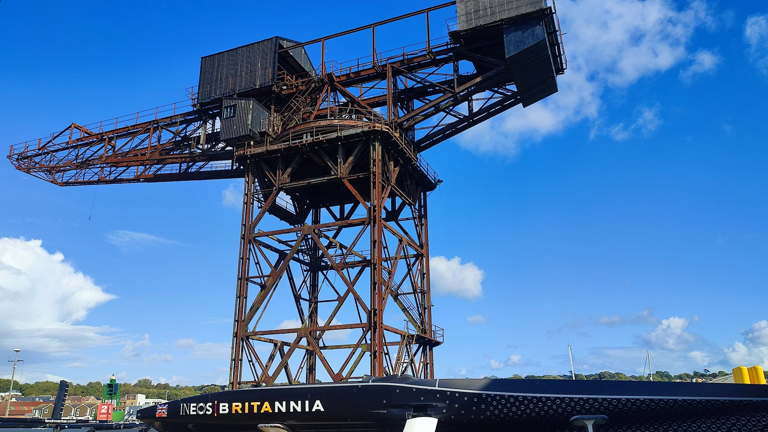 An old, rusted industrial crane with a large arm extending horizontally, situated on a waterfront with boats and a town in the background.