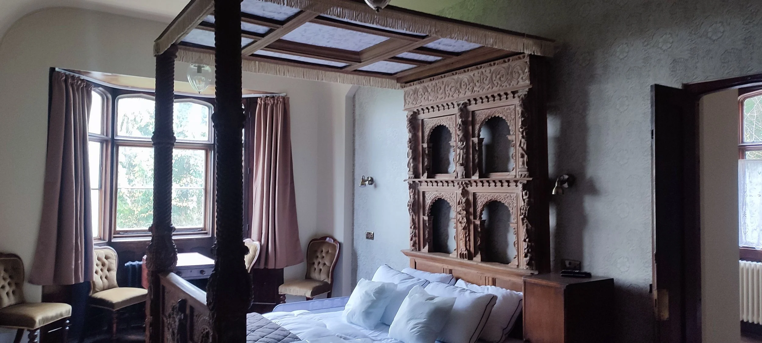 A bedroom with a four-poster bed, ornate carved wooden headboard, and a window with pink curtains.