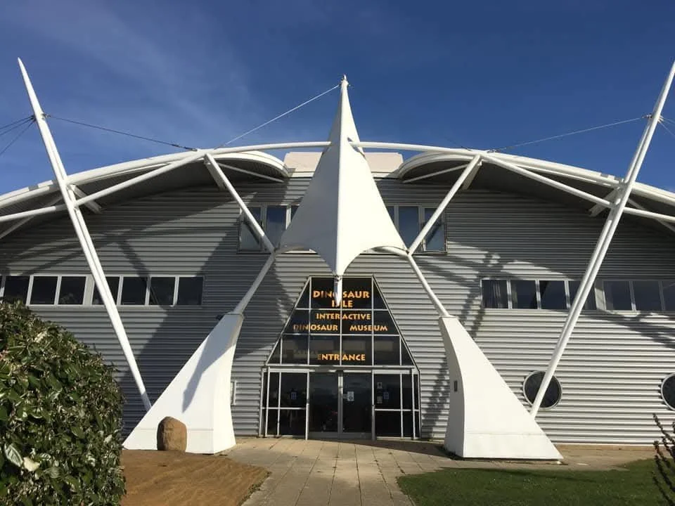 Exterior view of the Dinosaur Discovery Museum with a modern architectural design featuring large white structural elements and a metallic facade under a blue sky.