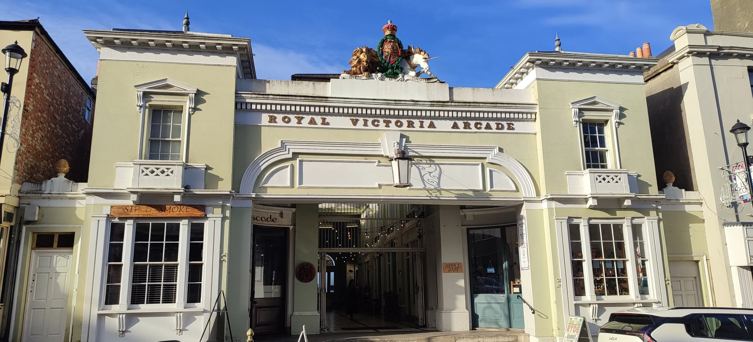 Historic building with the sign 'Royal Victoria Arcade' on top, featuring decorative elements including a lion and a horse statue, with storefronts and a car parked in front.