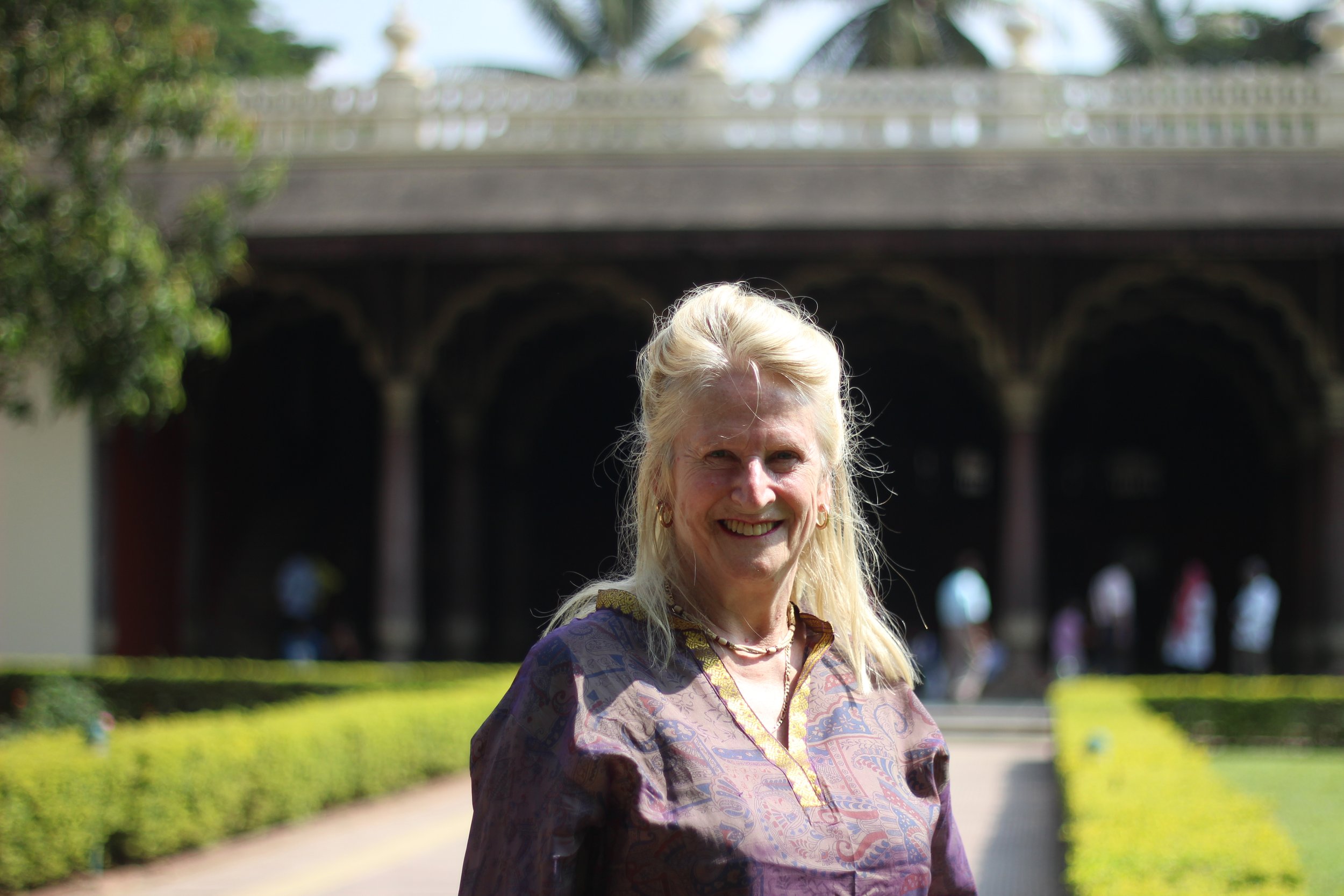 A smiling woman with long blonde hair standing outdoors in front of a dark bridge, with green bushes and trees on either side.