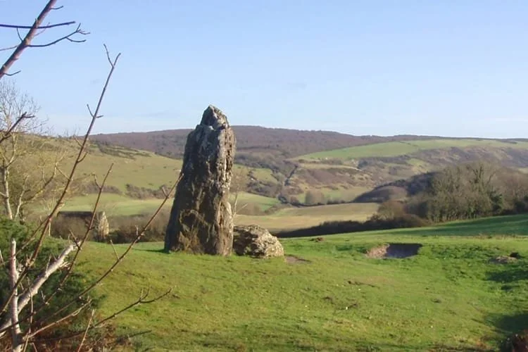 A large upright stone standing on a grassy landscape with rolling hills in the background.