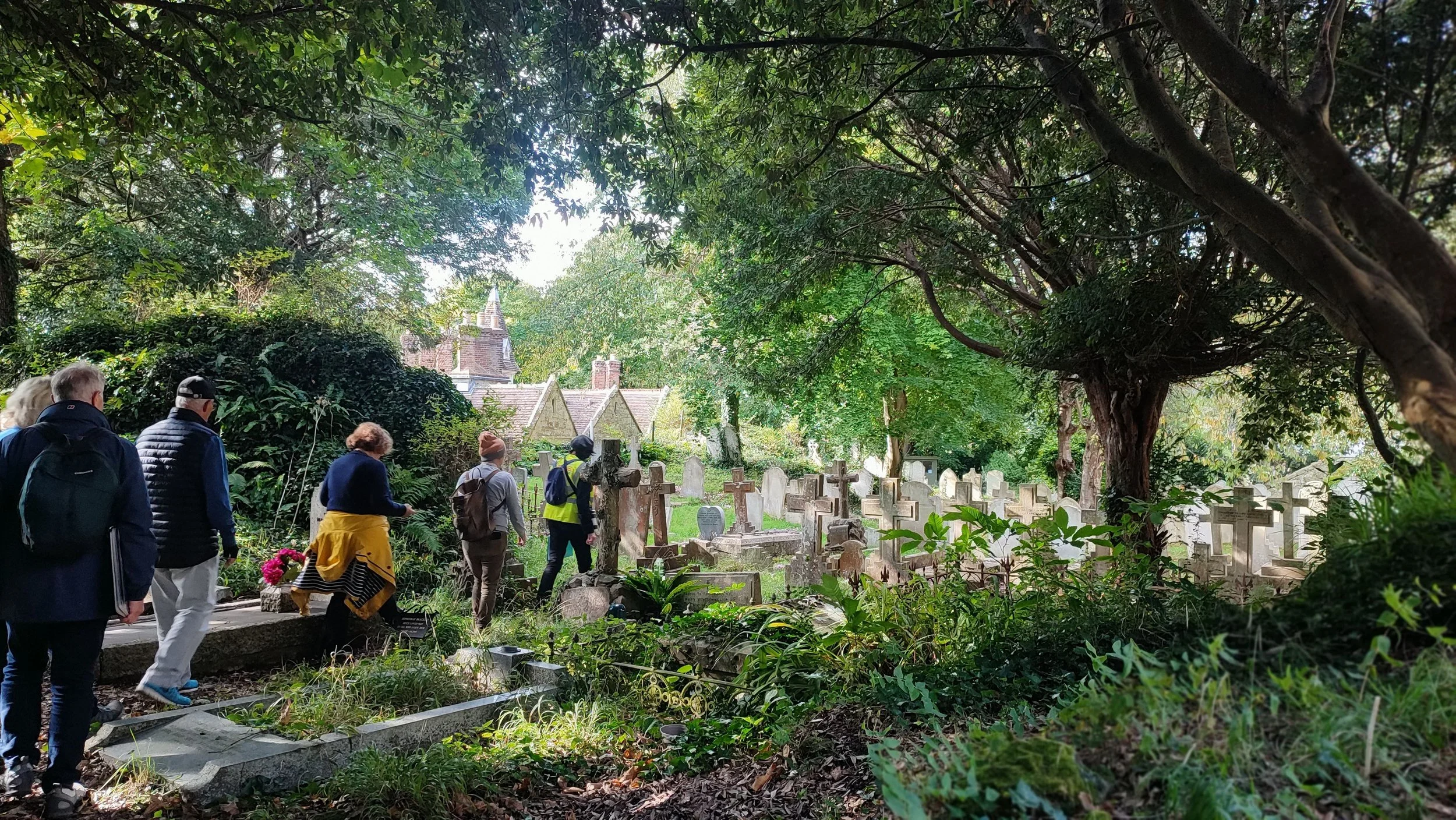 Group of people walking through an old cemetery with many stone and wooden grave markers surrounded by trees and greenery.