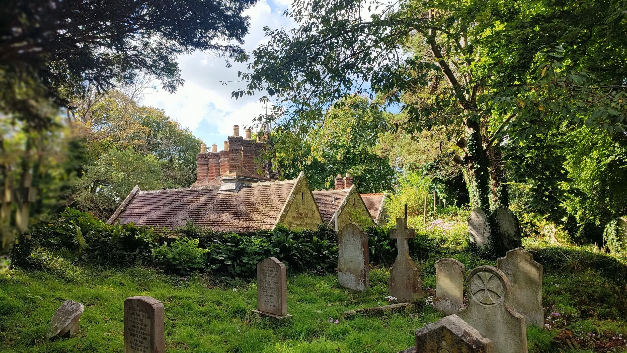 A peaceful old graveyard with various tombstones surrounded by lush green trees and foliage, and old houses with brick chimneys in the background.