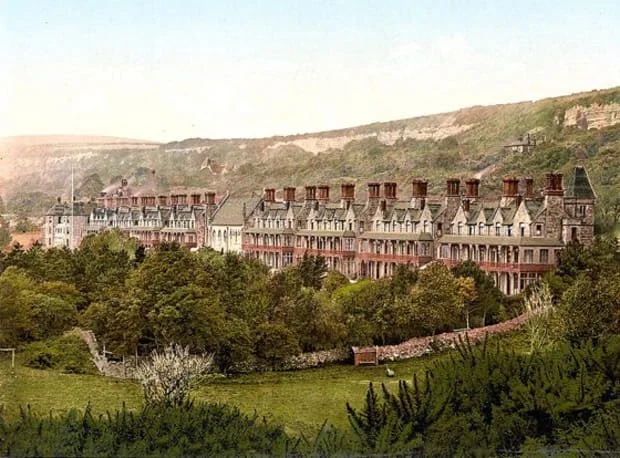 A row of large, historic buildings with multiple chimneys, set against a lush, green landscape with trees and hills in the background.