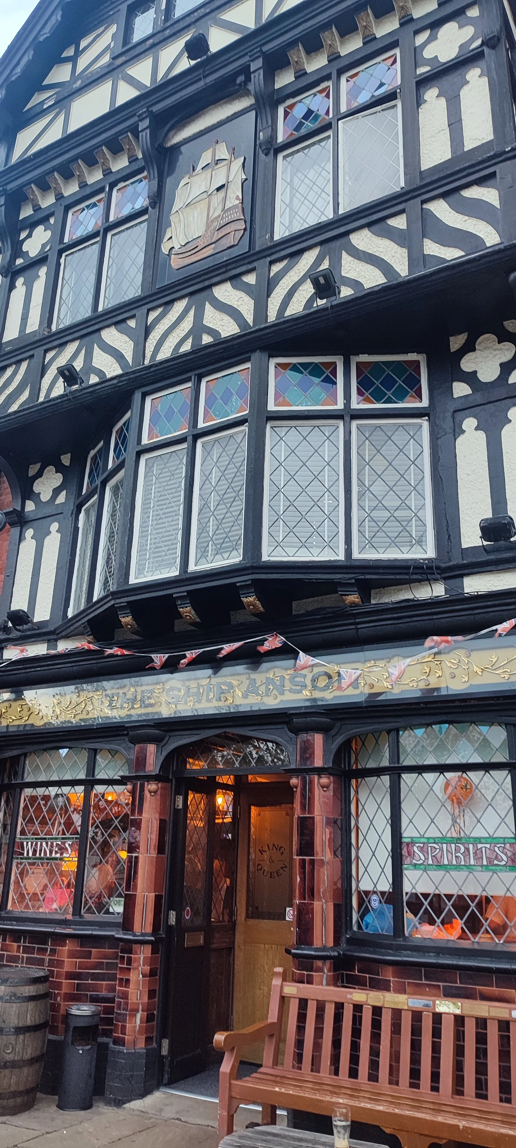 A building with black and white Tudor-style facade, stained glass windows, a ship decoration, and a pub entrance called 'The Ship & Queen' with outdoor seating.