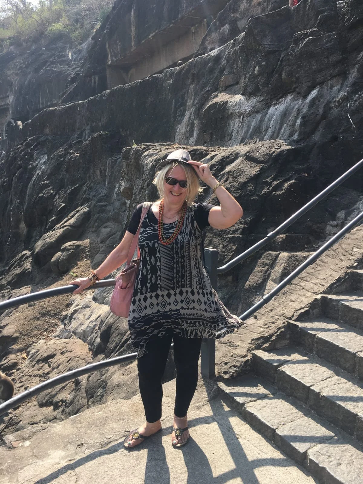 A woman with blonde hair wearing sunglasses, a black and white patterned dress, black leggings, and sandals, smiling while holding on to a metal handrail on stone stairs outdoors with rocky terrain and stone ledge behind her.
