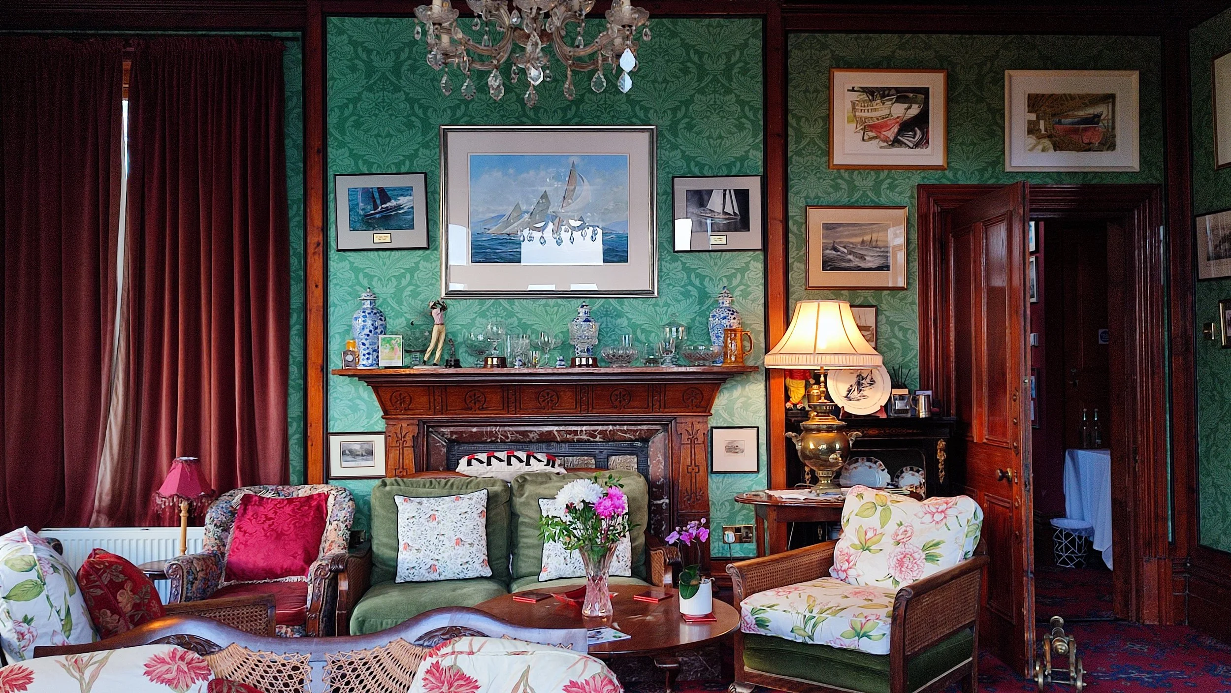 Living room with green floral wallpaper, dark wood trim, and red curtains. Features antique-style furniture, floral and velvet cushions, a wooden fireplace mantel with decorative vases, artwork, and a large mirror. A chandelier hangs from the ceiling, and there are lamps and flowers on the tables.