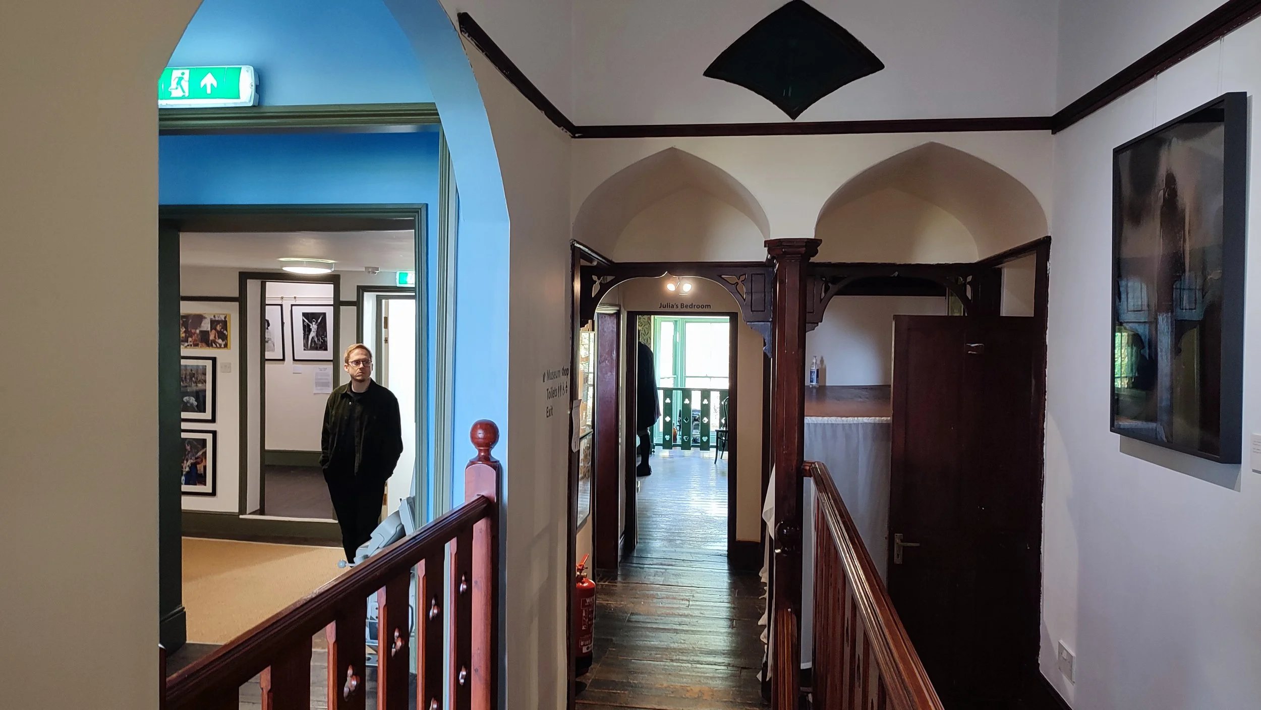 Interior of a museum with arched doorways, wooden railings, and framed photographs on the walls. A person is walking through a hallway in the background, and a fire extinguisher is visible on the floor.