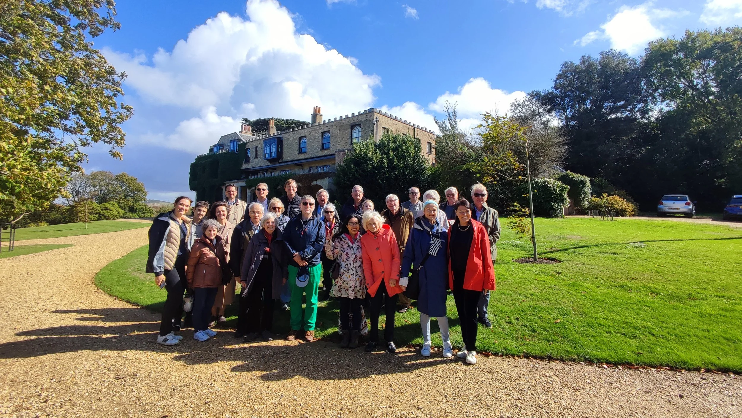 A group of people posing for a photo outdoors on a sunny day with a large historic building in the background, green lawns, trees, and a clear blue sky with some clouds.