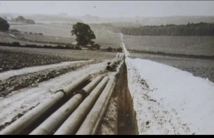 A dirt road with large pipes on the left and a white, snowy-looking field on the right, leading towards a tree and hills in the distance under a cloudy sky.