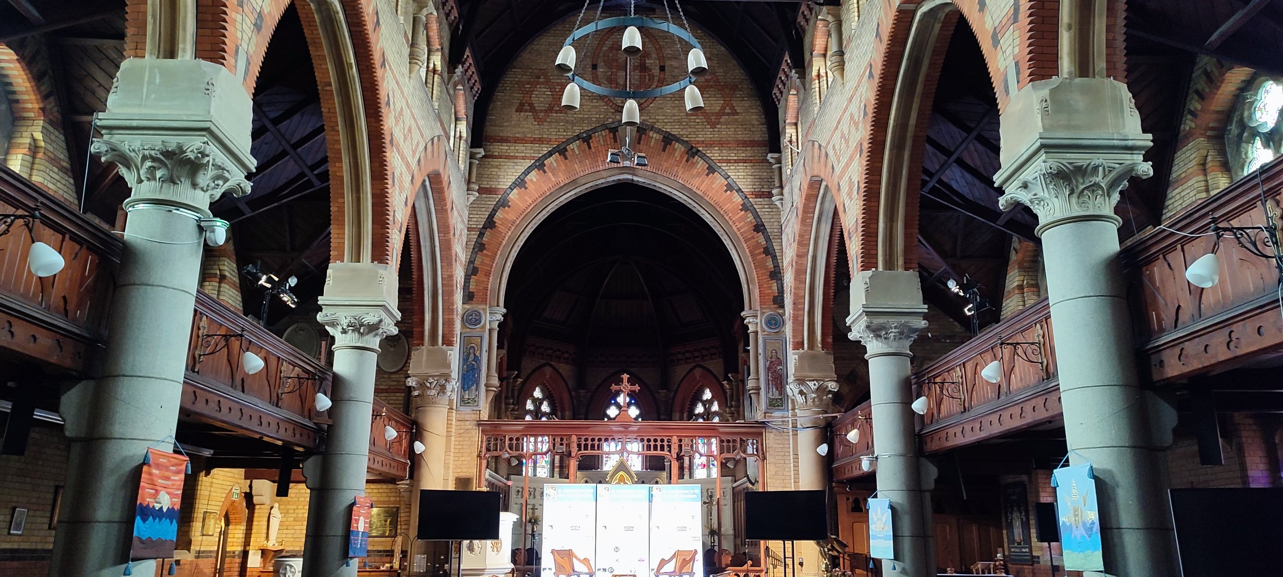 Interior of a church with tall stone columns, wooden balconies, stained glass windows, and religious artwork, with digital screens and banners on display.