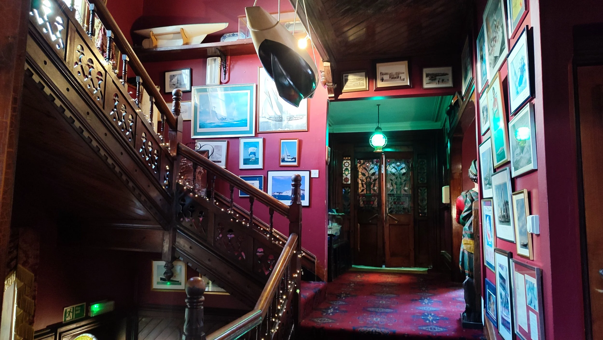 Interior of a cozy, vintage-style building with dark wooden staircase and walls decorated with framed pictures of boats and ships. There is a mannequin wearing a colorful dress on the right and a green hanging light fixture in the background.