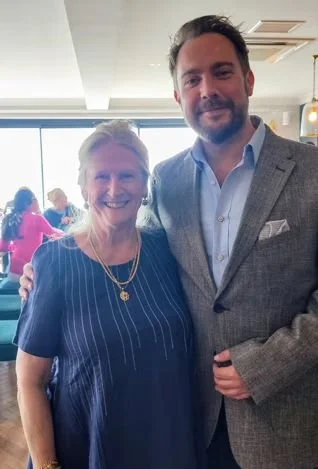 A woman and a man standing together indoors, smiling at the camera, with other people in the background.