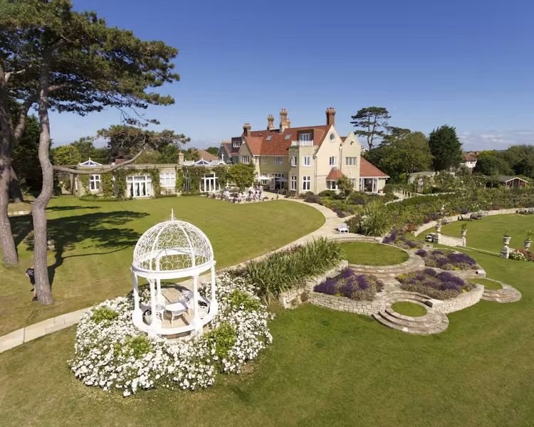 Large mansion with multiple stories, red tile roof, and extensive gardens with manicured lawns, stone pathways, and decorative flower beds, including a white gazebo in the foreground surrounded by blooming white flowers.