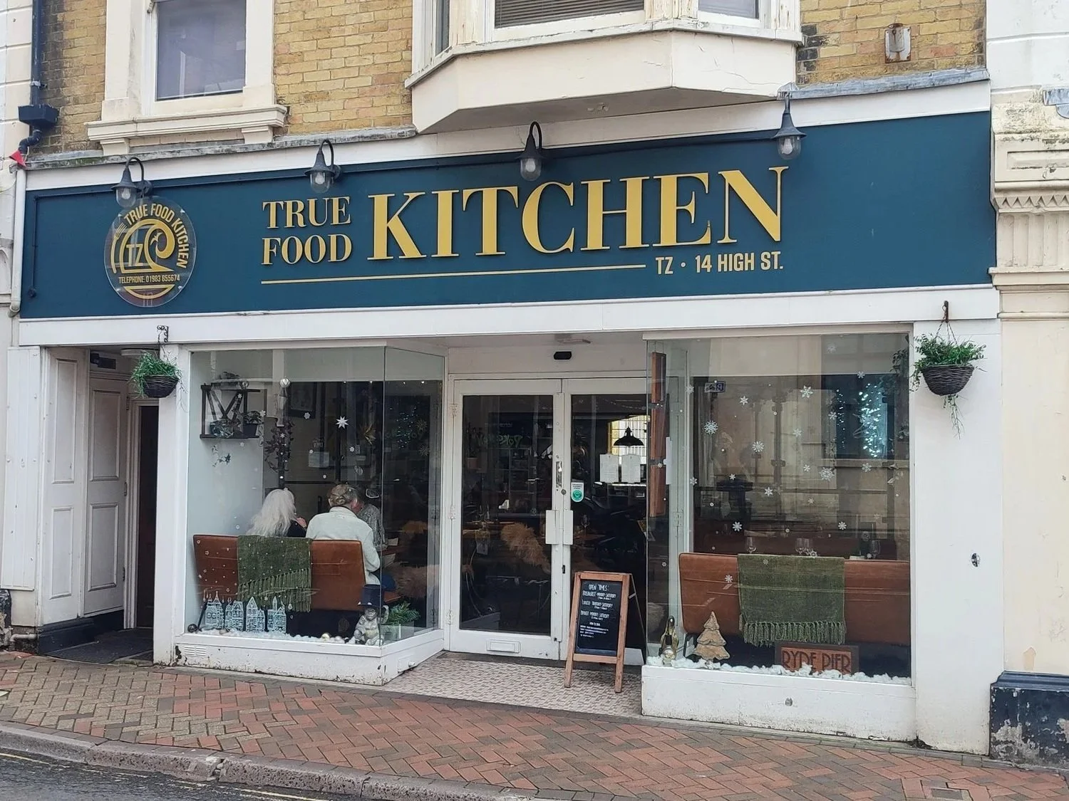 Facade of a restaurant called True Food Kitchen at 14 High Street, with a blue sign and glass windows showing customers inside, holiday decorations, and a chalkboard menu outside.