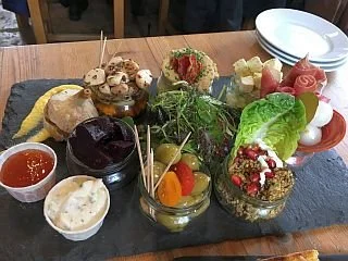 A platter with assorted appetizers and spreads, including small bowls of sauces, a variety of cheeses, vegetables, and garnishes, served on a black slate board.