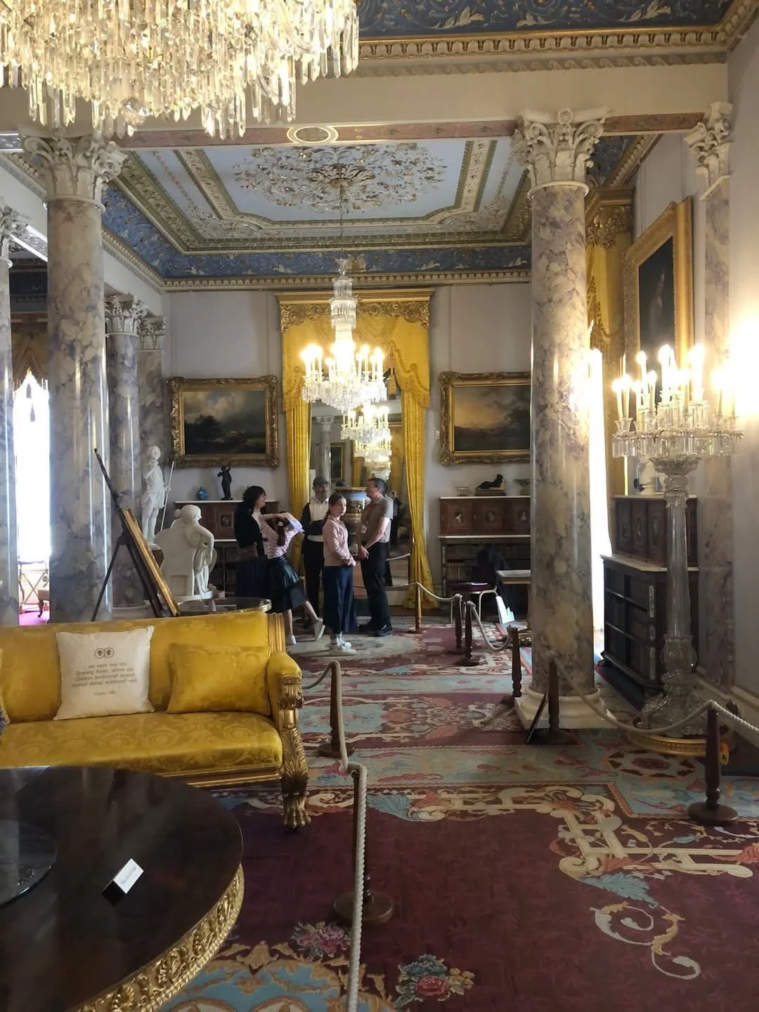 An opulent room in a historic building with detailed chandeliers, ornate moldings, marble columns, and classic paintings. Several people are gathered in conversation near a display area, with luxurious furniture and decorative elements throughout.