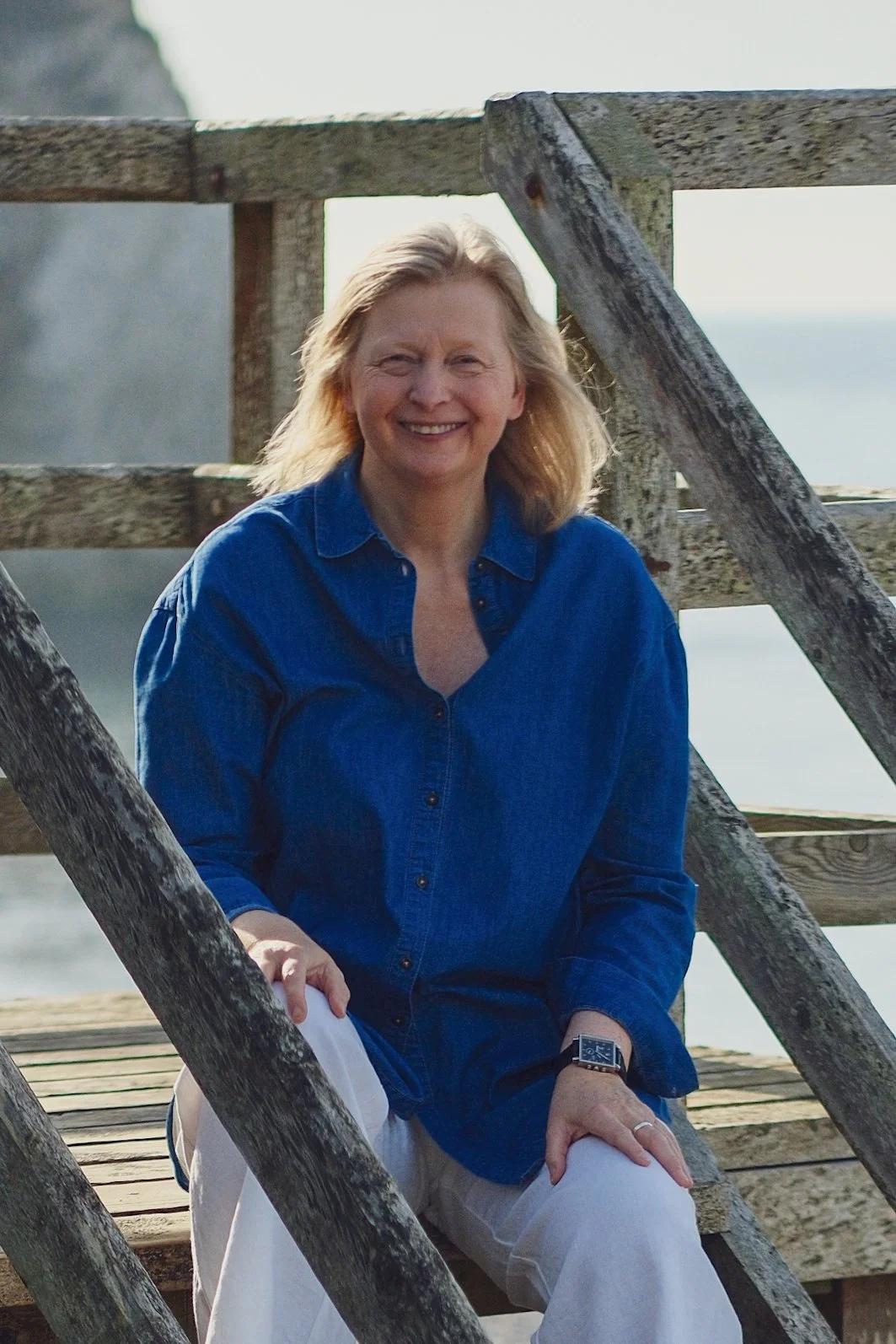 A woman with blonde hair wearing a blue denim shirt and white pants, smiling while sitting on a wooden structure outdoors near the water.