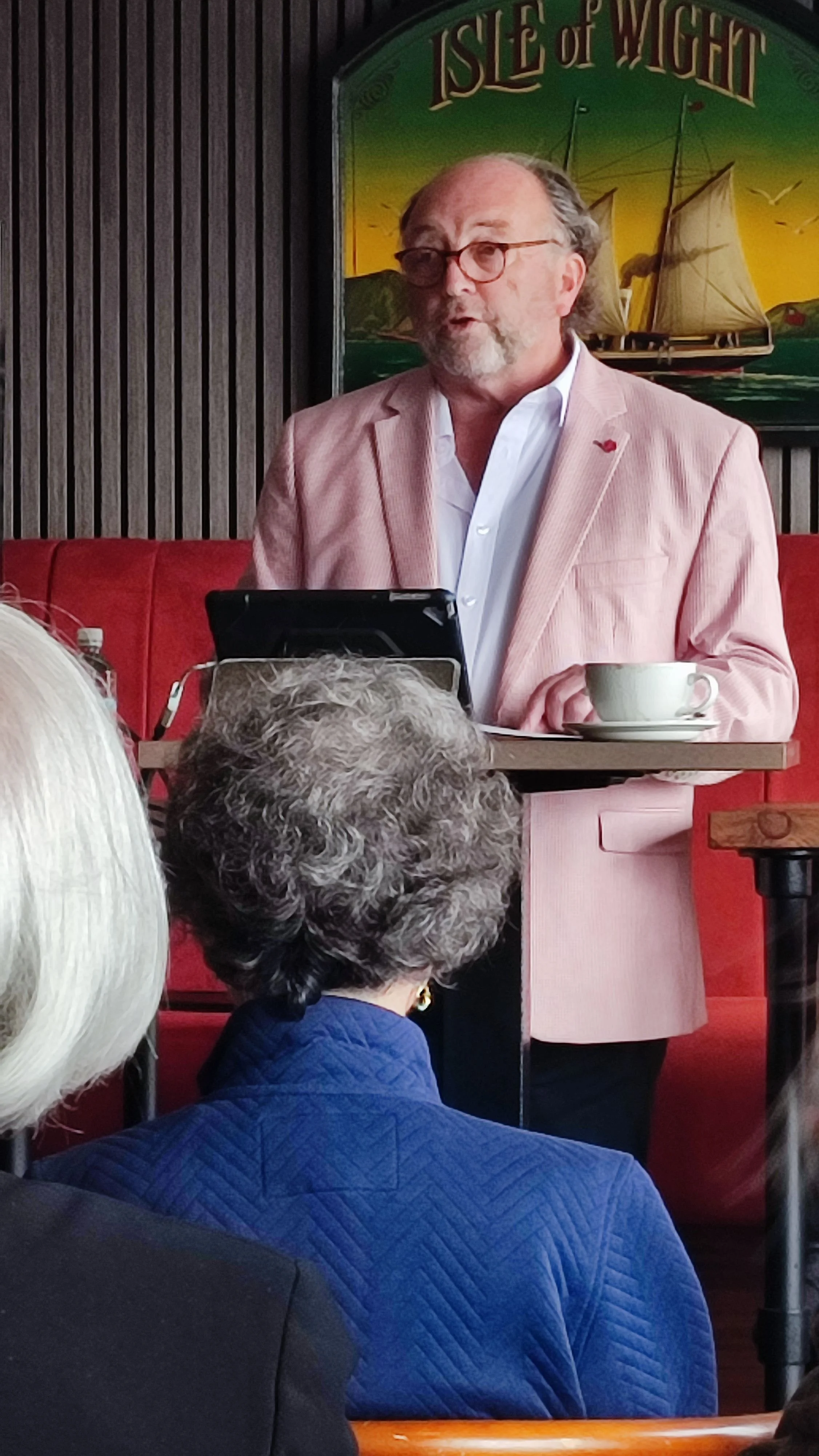 A man with glasses, a beard, and gray hair speaking at a podium in a restaurant or cafe. He is wearing a light pink blazer and white shirt, with a cup of coffee or tea on the table beside him. Behind him is a colorful sign that says 'Isle of Wight' with a painting of a sailing ship.