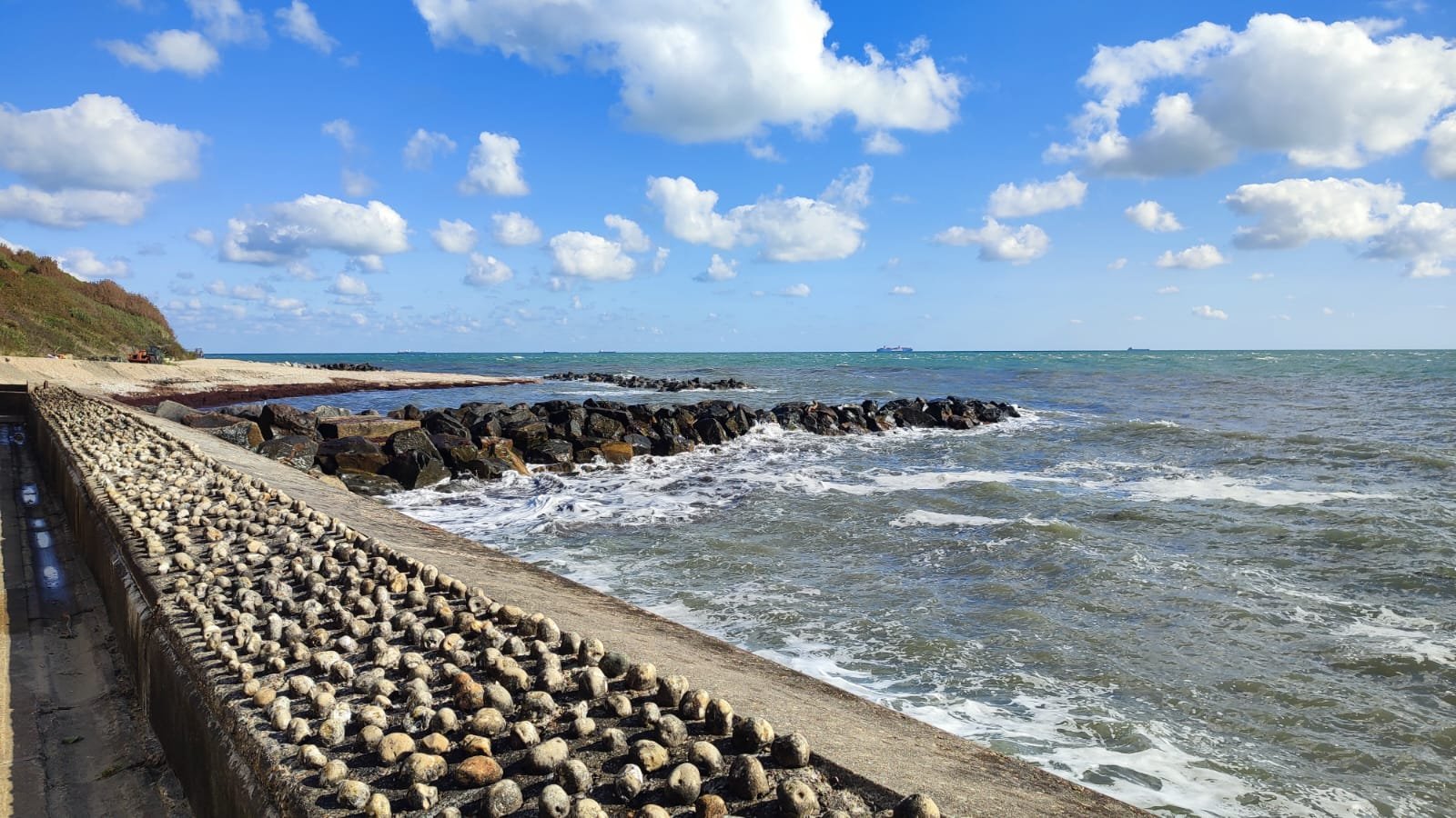 Sea waves crash against a rocky barrier along a beach with a distant ship on the horizon and a partly cloudy blue sky.
