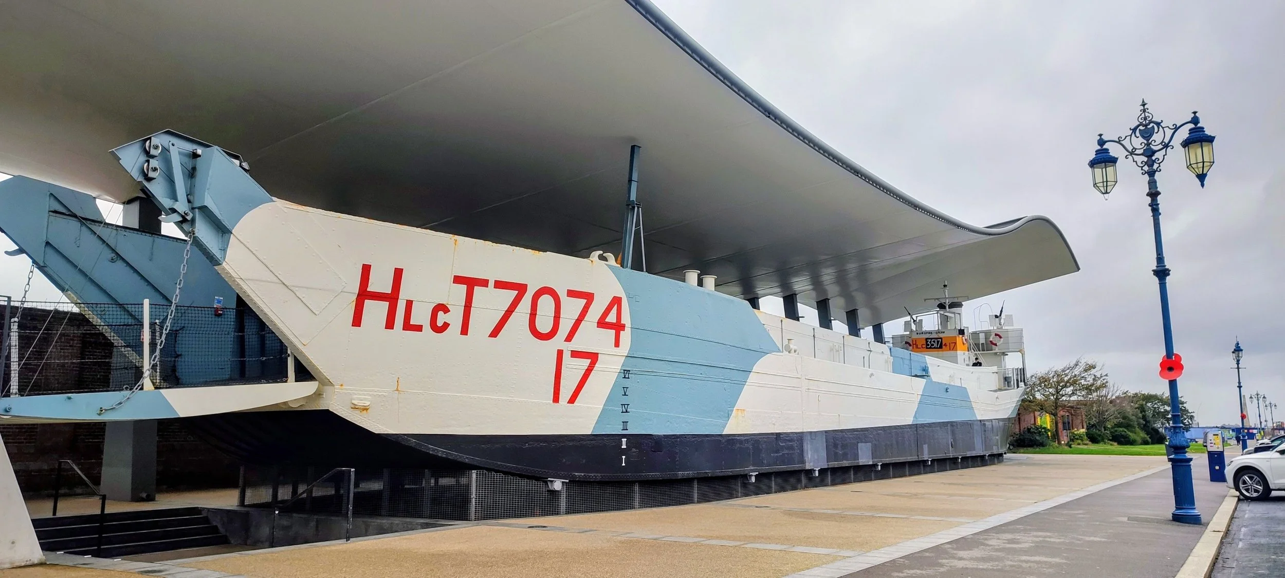 An aircraft carrier ship on display outdoors, featuring a large white and blue hull with the red identification number 'HLC 7074' and the number 17. The ship has a distinctive curved superstructure and is surrounded by a concrete walkway with street lamps and cars parked nearby.