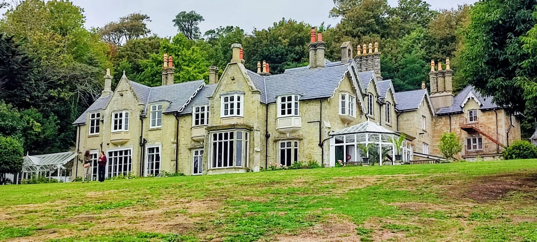 A large historic mansion with stone walls and many chimneys, surrounded by trees and greenery, with two people standing near a greenhouse on the lawn.