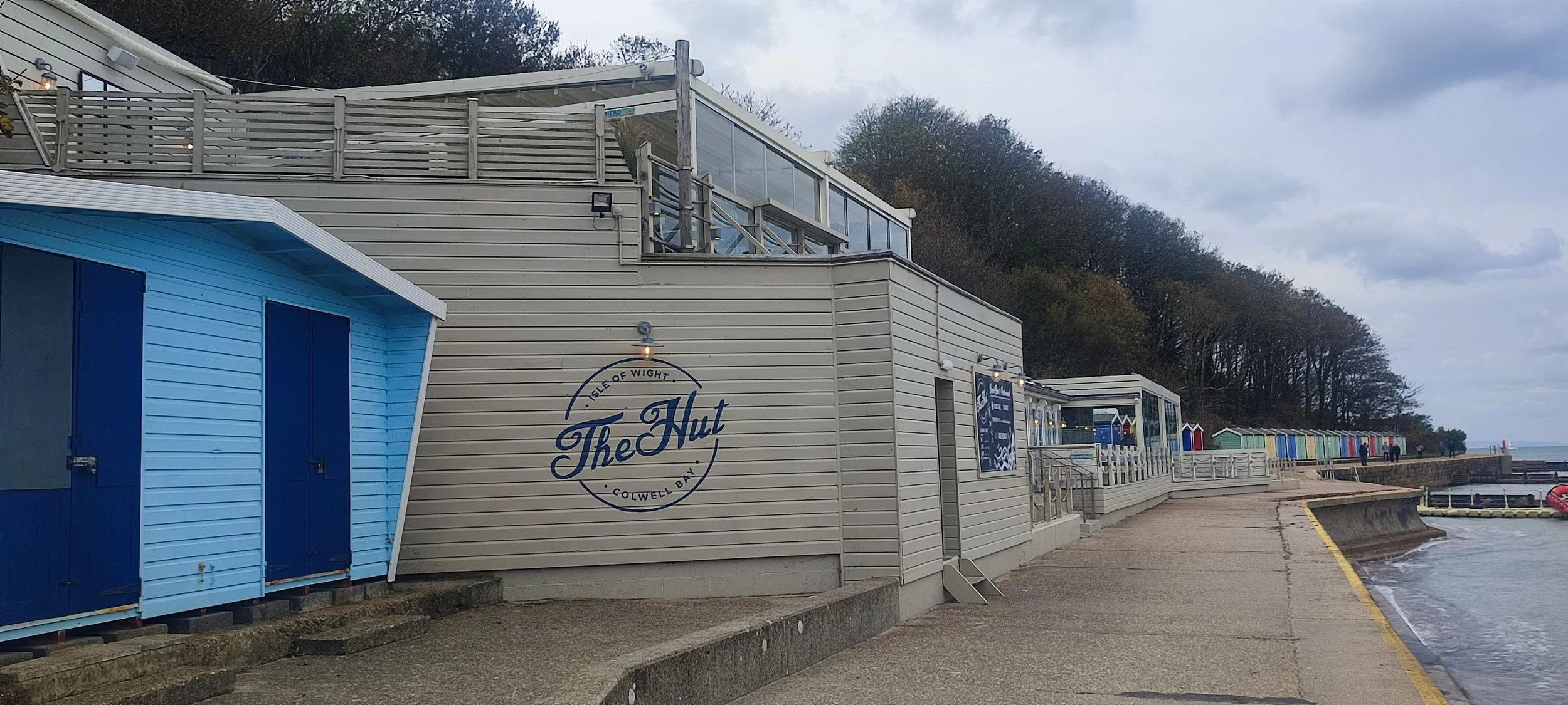 A seaside promenade with colorful beach huts, a building with a sign saying 'The Hut' on the Isle of Wight, Calwell Bay, with a cloudy sky and trees on a hillside in the background.
