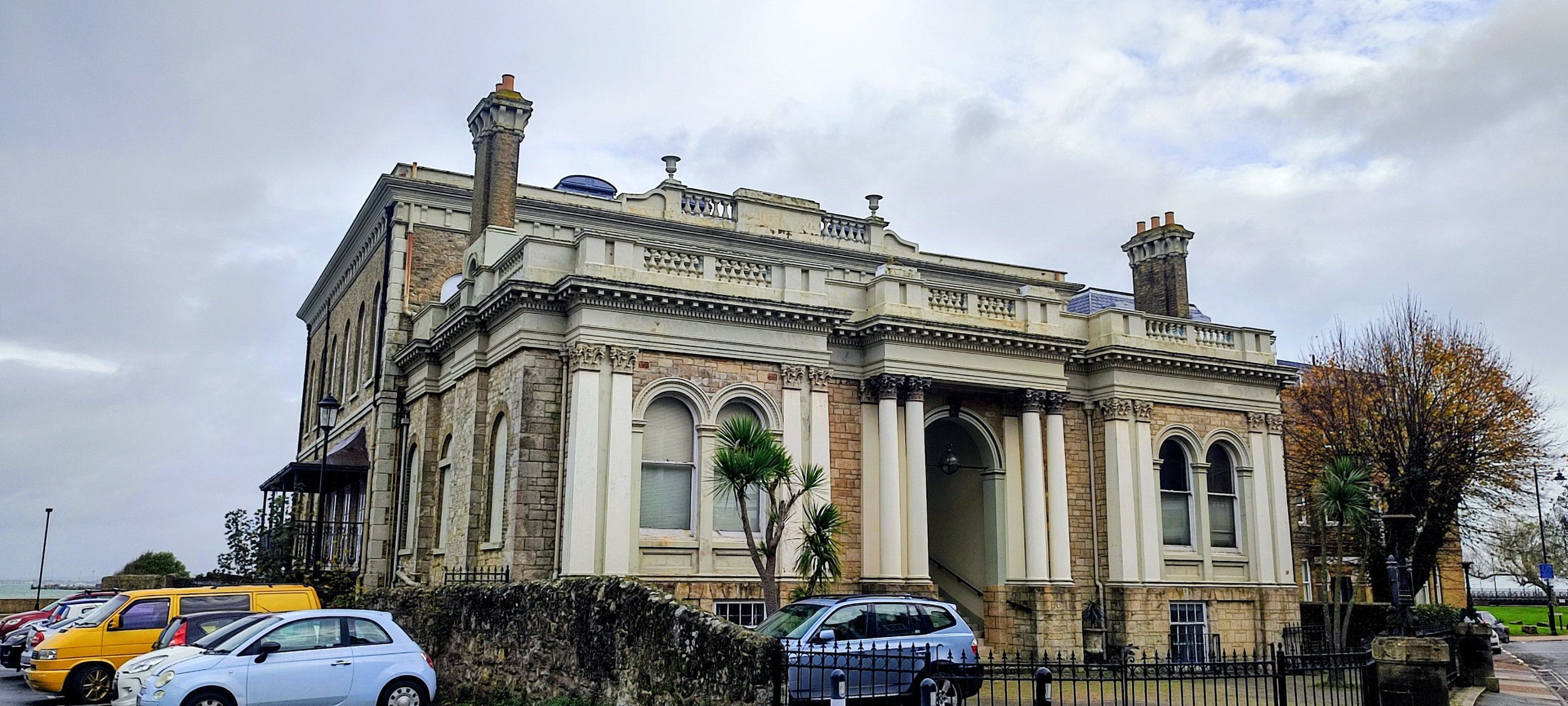 Large historic mansion with classical columns, stone and brick facade, surrounded by parking and trees, overcast sky