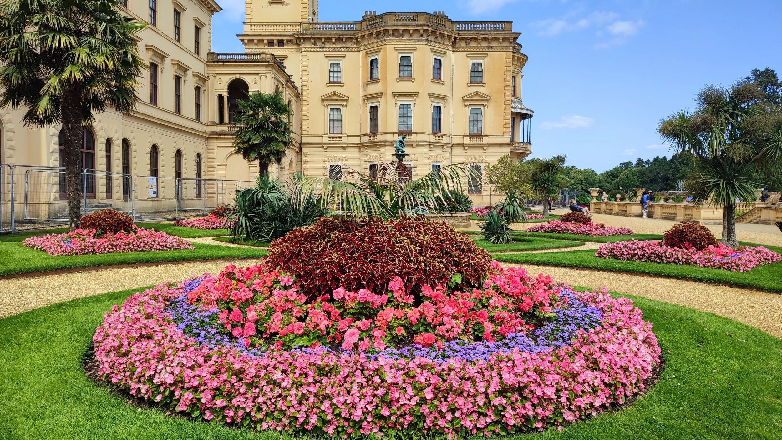 A historic yellow Victorian-style building with ornate architectural details, surrounded by a well-maintained garden with colorful flowers, green lawns, and palm trees.